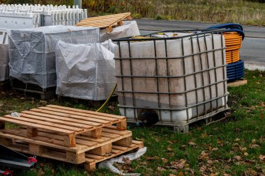 Konteynırlar ve paletler yol yakınındaki bir inşaat alanında düzenlenir. Bölge çimenler ve ağaçlarla çevrilidir. Sahne, çeşitli materyallerle çalışma hazırlıklarını gösteriyor.