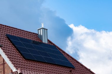 Güneş panelleri, açık mavi gökyüzünün altında bazı bulutlarla birlikte güneş ışığını yansıtan kahverengi kiremitli bir çatıya monte edilir. Bu kurulum bir evde yenilenebilir enerji kullanımını gösterir.