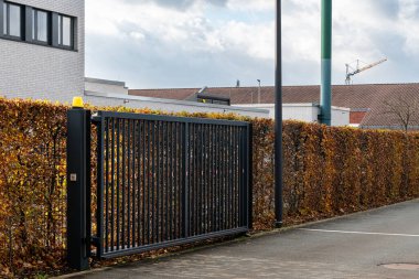 Şık metal bir kapı, kentsel bir ortamda düzgünce kırpılmış bir çitin yanında açılıyor. Sahne mimari ve yeşilliğin bir karışımını yakalıyor, güneşli bir günde iyi korunan bir açık hava alanı sergiliyor..