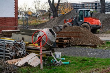 Yoğun bir inşaat sahasında çimento karıştırıcı ve yığınla yapı malzemesi görülüyor. Bir traktör, arka planda sonbahar ağaçları olan gri gökyüzünün altındaki toprak yığını üzerinde görülebilir..