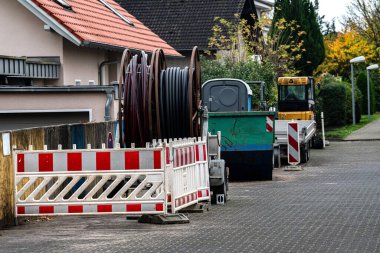 Mahalledeki bir sokakta ağır makineler ve inşaat bariyerleri görülüyor. Kablo makaraları yakına yerleştirilmiş, devam eden kamu hizmetlerini gösteriyor.