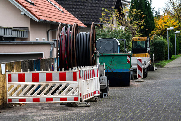 Heavy machinery and construction barriers are seen on a street in a residential neighborhood. Cable reels are placed nearby, indicating ongoing utility work.