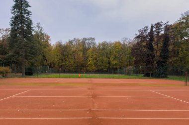 Kil tenis kortu boş, canlı sonbahar ağaçlarıyla çevrili. Sahne sükuneti yakalıyor, yapraklar renk değiştiriyor ve görünürde hiç oyuncu yok, huzurlu bir gün için mükemmel..