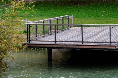 Suyun üzerinde, yemyeşil çimlerle çevrili ahşap bir izleme platformu duruyor. Nehir kıyısı boyunca uzanan ağaçlar, ziyaretçilere gölge ve güzellik sunuyor.. 
