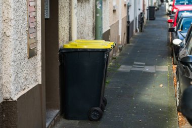 Sarı kapaklı siyah bir çöp tenekesi dar bir şehir kaldırımında bir binaya dayanıyor. Park halindeki araçlar caddelerde kuyruk oluşturuyor ve olay yeri gün içinde tipik bir şehir ortamını yakalıyor..