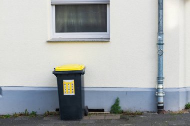 Sarı kapaklı siyah bir çöp kutusu düz bir bina duvarının yanında duruyor. Üssün yakınında küçük bir çimen parçası görülebiliyor ve sahne gün ışığında çekiliyor..