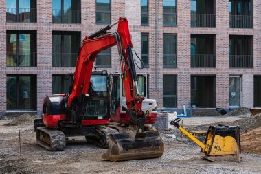Kırmızı kazıcı ve şantiyede daha küçük bir sarı sıkıştırıcı. Arka planda gelişmekte olan yeni tuğla binalar yer alıyor. Gün doğumu açık görüş mesafesinde..