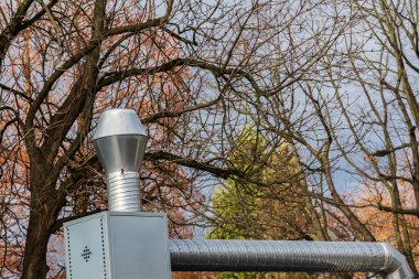 Gümüş bir baca, yapraksız ağaçlarla çevrili gri bir gökyüzüne çarpar..