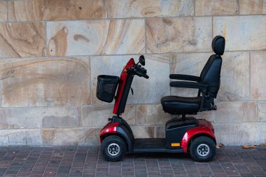 Kırmızı, yaşlı, hareketli bir scooter, desenli taş bir duvarın yanında duruyor. Mobilet kullanıma hazır, rahat bir koltuk ve sepet konfor için..