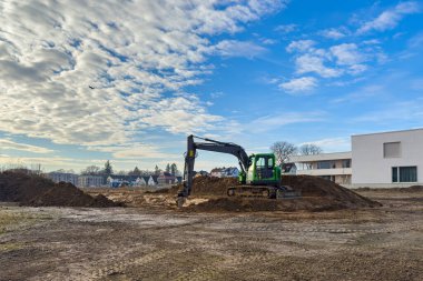 Bir kazıcı bulutlu bir gökyüzünün altındaki bir inşaat sahasında toprak yığınını kazıyor. Manzara, gün boyunca arka planda binalar olan temiz bir zemin gösteriyor. Bir kazıcı bir tümseği kazıyor.