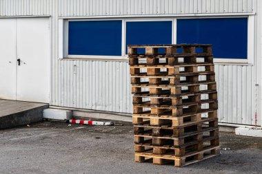 Binanın dışında bir yığın ahşap palet duruyor. Paletler beyaz bir duvar ve mavi pencerelerin yanındaki gri asfaltta düzgünce düzenlenmiş. Ayarlar gündüz vakti ticari bir alanda yapılıyor..