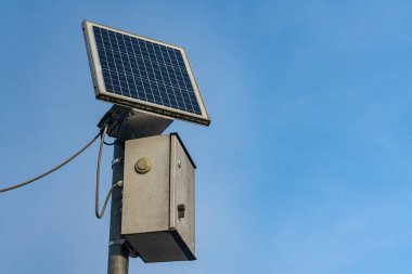Bir güneş paneli metal bir kutuya bağlanmış bir kontrol kutusuyla monte edilir. Arka plan bulutsuz açık mavi bir gökyüzünü gösteriyor. Güneş ışıl ışıl parlıyor. Gündüzü gösteriyor..