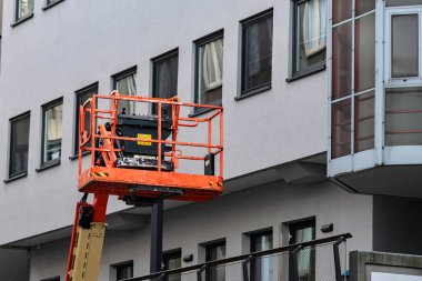 İşçiler tamirat için bir binanın üst katlarına ulaşmak için bir inşaat asansörü kullanıyorlar. Bu sahne yoğun bir şehir merkezinde gündüz vakti geçmektedir..