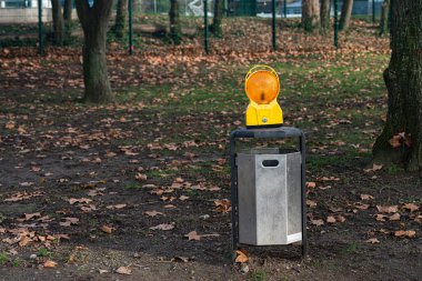 Düşen yapraklarla çevrili bir parktaki çöp kutusunun üstünde bir trafik lambası duruyor. Sahne arka planda ağaçları ve kuru yapraklarla kaplı zemini gösteriyor..