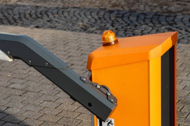 Şehir merkezinde bir trafik bariyeri sistemi görülüyor. Bariyer kolu aşağıda ve tepesinde bir uyarı ışığı yanıp sönüyor. Yerde kaldırım taşları var ve yıpranma izleri var..