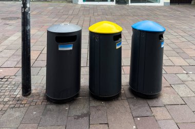 Şehir caddesinde üç çöp bidonu yan yana duruyor. Kutular siyah, bir sarı kapağı ve bir mavi kapağı var. Bölgede tuğlalar ve arkalarında bir dükkan var..