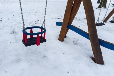 Bir oyun parkı boş salıncaklar ve karla kaplı tahterevalliler gösterir. Ekipmanların parlak renkleri beyaz zemine karşı duruyor. Bugün oyun oynayan çocuklar yok..