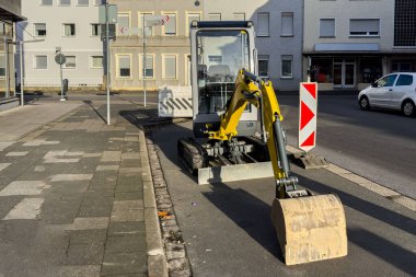 Küçük bir kazıcı bir kentsel alanda yolun kenarında dinleniyor. Yakınlarda sokak tabelaları ve park edilmiş arabalar var. Sahne gündüz açık gökyüzü ile çekiliyor..
