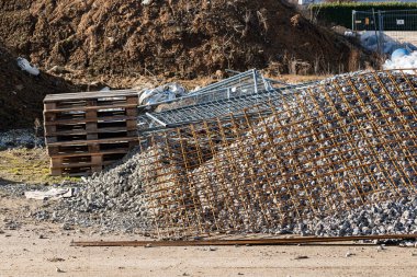 İnşaat sahasında yerde duran ahşap paletler, çakıl yığınları ve metal inşaat demirleri yer alıyor. Yakınlarda, gün ışığında toprak yığınları var..