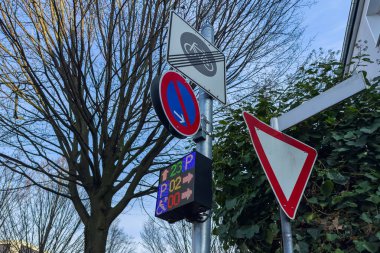 Dijital park ekranı olan bir şehir bölgesinde trafik işaretleri var. Açık mavi gökyüzü saatin gündüz olduğunu gösteriyor ve yakınlardaki ağaçların yaprakları yok..