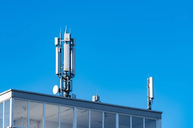 İki telekomünikasyon kulesi açık mavi bir gökyüzünün altında bir binanın tepesinde duruyor. Kuleler, bugün şehirlerde kullanılan teknolojiyi gösteren mobil ağlar ve sinyaller için kullanılan ekipmanlardır..