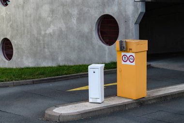 Bir otoparkta bir caddenin kenarında iki kutu standı var. Biri sarı, diğeri ise beyaz renklidir. Bölgede gri bir duvar ve arka planda yeşil çimenler var..