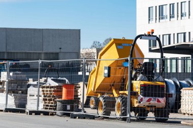 Sarı bir damperli kamyon bir inşaat sahasına park edilmiş. Etrafında inşaat malzemeleri ve bir çit yığını var. Bölge, açık bir günde yoğun çalışma sürecini gösteriyor.