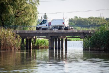 Yeşil kamışlı küçük bir nehrin üzerindeki beton köprüde beyaz tanker kamyonu. Gri beton sütunlu yol köprüsü.