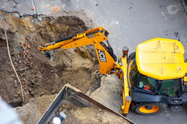 Ağır makineler, bir kentsel bölgedeki ısıtma sistemini onarmak için toprağı kazıyor. Çalışma alanı toprak ve inşaat malzemelerinin taşındığını gösteriyor..