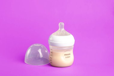 Bir Philips Avent süt şişesi parlak mor bir yüzeye oturur. Şişe berrak bir kapakla kapalı ve süt içeriyor. Bu sahne modern bir bebek beslenme aksesuarı yakalar..