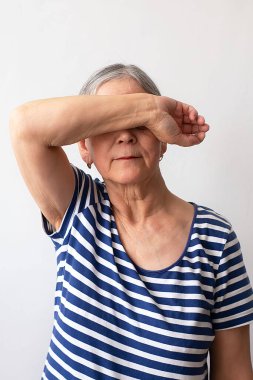 Yaşlı bir kadın, 60 'lı yaşlarda beyaz saçlı, çizgili beyaz ve mavi elbiseli, elini beyaz arka planda tutan gözlerini kapatan bir kadın..