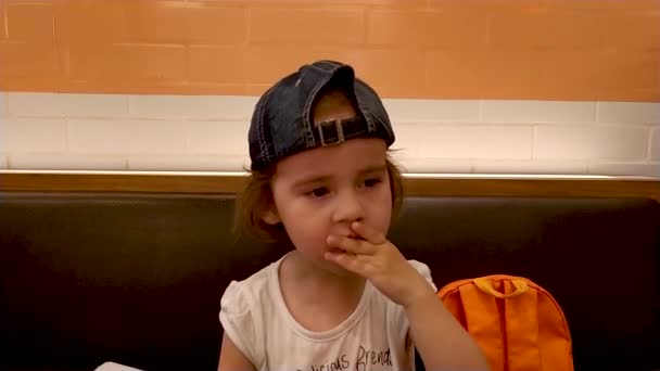 Ребенок сидит в кафе. Маленькая девочка в бейсболке ест фаст-фуд — стоковое видео