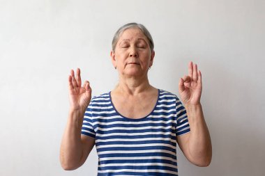 Yaşlı bir kadın, 60 'lı yaşlarda, çizgili beyaz ve mavi elbiseli gri saçlı bir kadın, parmaklarıyla meditasyon hareketi yaparken rahatlıyor. Yoga konsepti