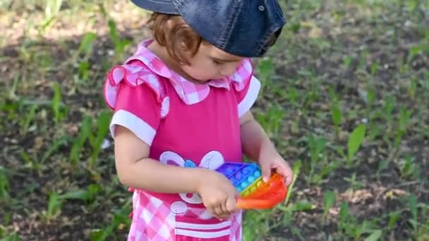 Симпатичная маленькая девочка в красном платье и бейсболка играть в природе с Fidget поп Это, гибкие сенсорные игрища с толкать пузырь — стоковое видео