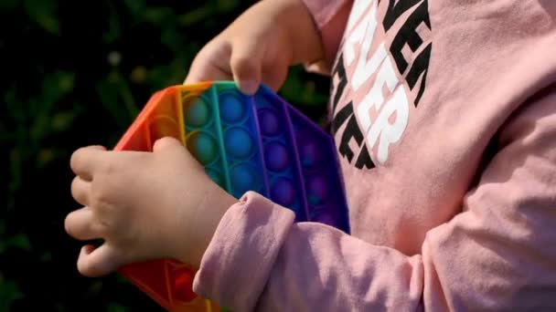 Uma criança brinca na rua com um brinquedo sensorial pop it. A menina pressiona colorido arco-íris macio bolhas de silicone macio. Alívio de estresse e ansiedade. Jogo de moda agitação — Vídeo de Stock