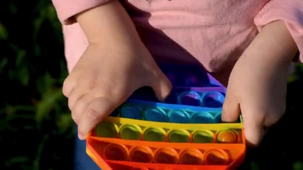 Un bambino gioca per strada con un giocattolo sensoriale pop it. La ragazza preme colorate bolle di silicone morbido arcobaleno. Alleviare lo stress e l'ansia. Gioco di agitazione di moda — Video Stock