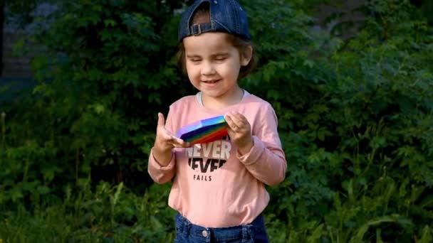 Un bambino gioca per strada con un giocattolo sensoriale pop it. La ragazza preme colorate bolle di silicone morbido arcobaleno. Alleviare lo stress e l'ansia. Gioco di agitazione di moda — Video Stock