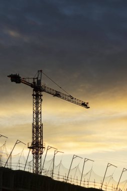 Bir vinç silueti ve inşaat halindeki bir binanın tepesi. Gün batımında veya gün doğumunda sarı ve mavi gökyüzü.