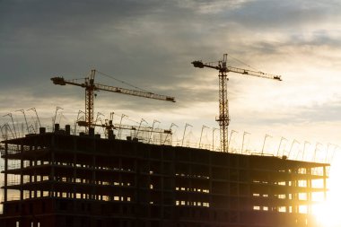 İki vinç silueti ve inşaat halindeki bir bina gökyüzünde bulutlar ve gün batımı veya gün doğumu ışığı.