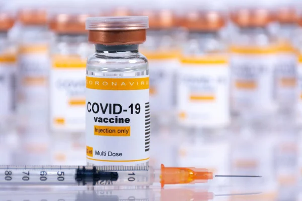 Coronavirus aşı şişesi ve beyaz bir masanın üzerinde yansımaları ve odak noktasının dışında başka şişeler olan bir şırınga. Laboratuvar ya da sağlık tesisi kavramı