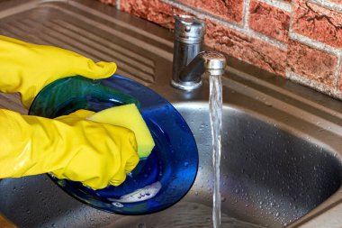 Lastik eldivenli kadın bulaşık ve tabakları süngerle yıkıyor mutfaktaki metal lavaboda..