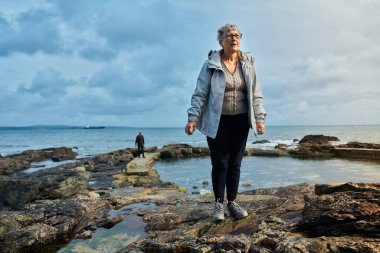 Son sınıf öğrencisi bir kadın sonbaharda bir plajın kayalıklarında duruyor.