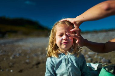 Sonbaharda plajda bir anaokulu öğrencisi yüzüne güneş kremi sürüyor.