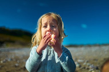 Bir anaokulu öğrencisi güneşli bir sonbahar gününde sahilde elma yiyor.