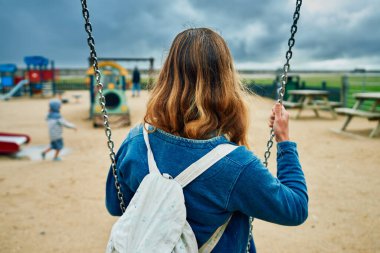 Bir anne salıncakta oturmuş anaokulunun parkta oynamasını seyrediyor.