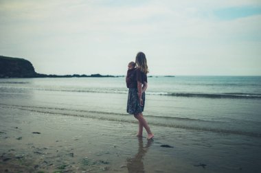 Genç bir anne, bebeği askıda kumsalda yürüyor.