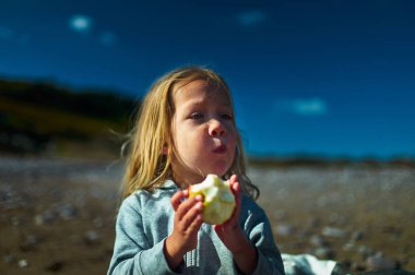 Bir anaokulu öğrencisi güneşli bir sonbahar gününde sahilde elma yiyor.
