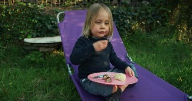 Küçük çocuk dışarıda karyolada otururken yemek yiyor.