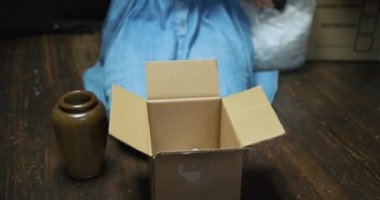 Kadın elinde köpük parçalarıyla karton kutuda vazo paketliyor.