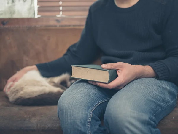 Young man sitting on a a sofa with a cat and a big book — Stock Photo, Image